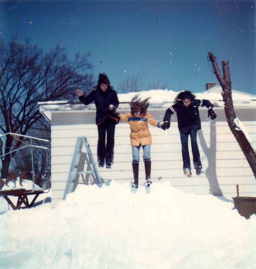 Three people jump off a snowy roof, holding hands, under a clear blue sky with bare trees in the background.