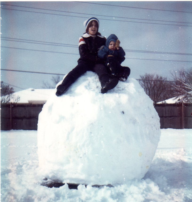 Two children sit atop a large snowball, wearing winter clothing, in a snow-covered yard with houses and trees in the background.