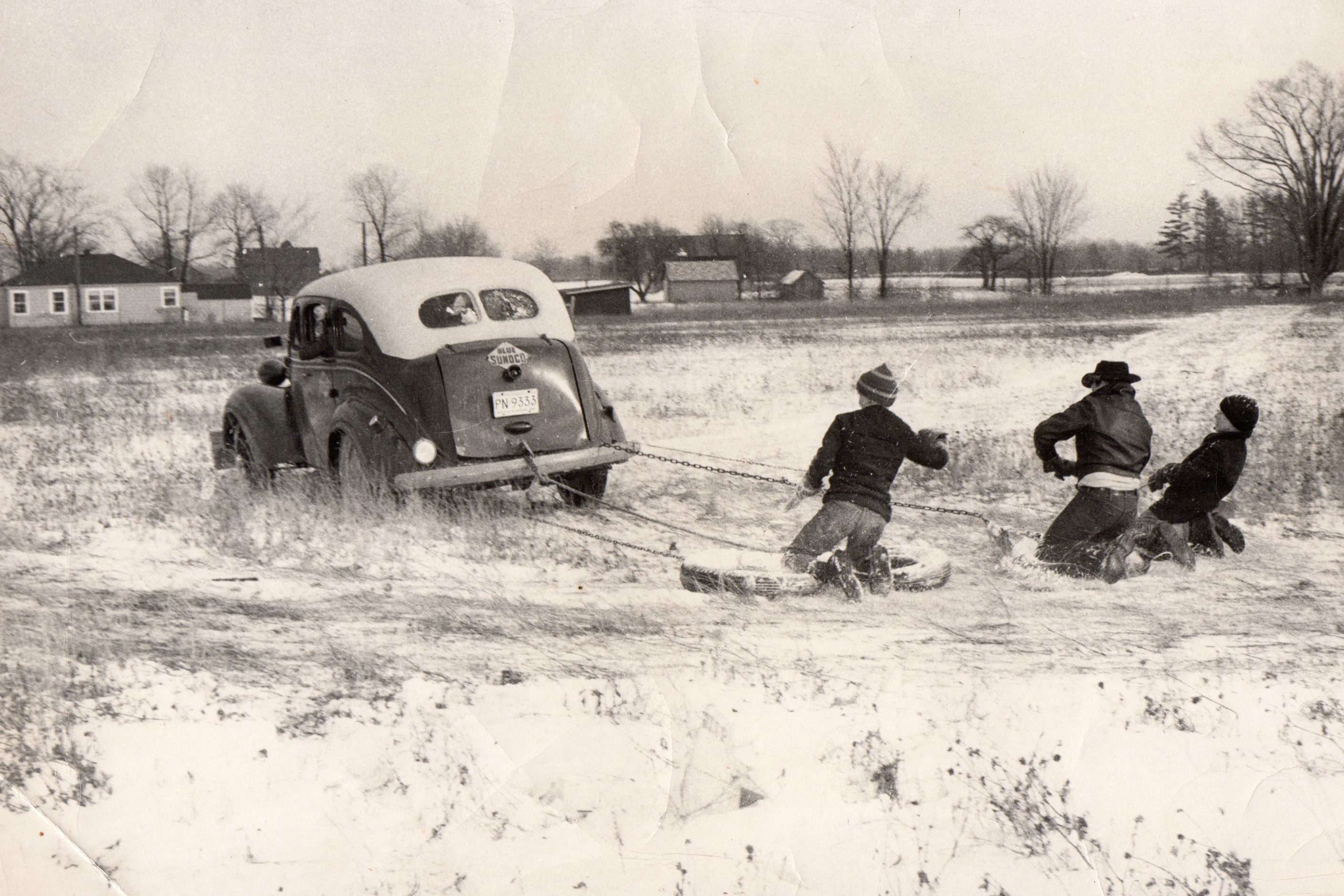 Car pulls three people on sleds, snowy field in background.