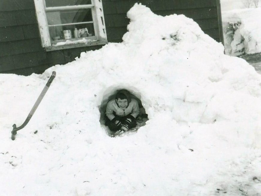 Person crouching inside a small snow cave near a house, with a shovel leaning against the snowy mound.