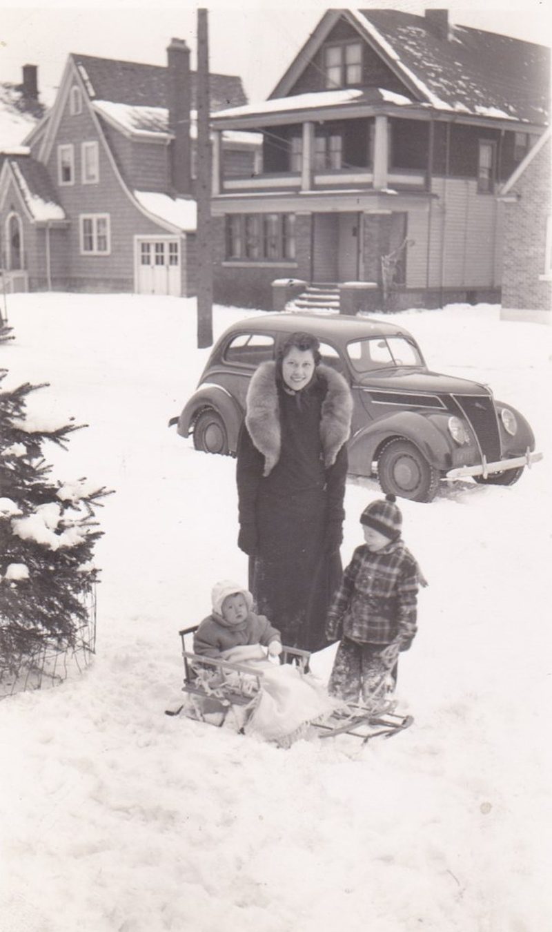 Woman stands with two children and a sled in snowy street, vintage car and houses in background.