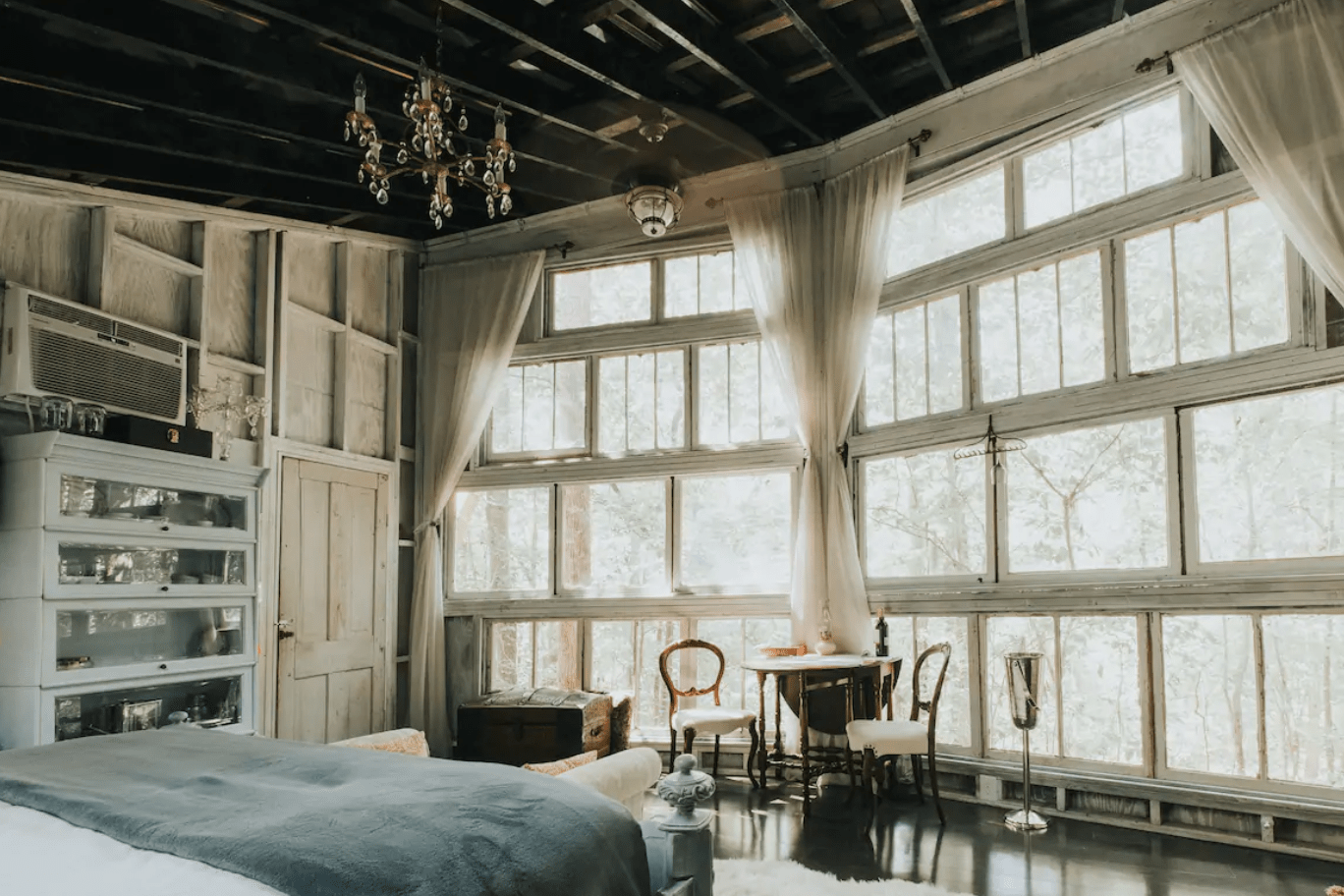 Bedroom features large windows, sheer curtains, and elegant furniture, including a chandelier and small table set, creating a vintage-style, airy ambience.