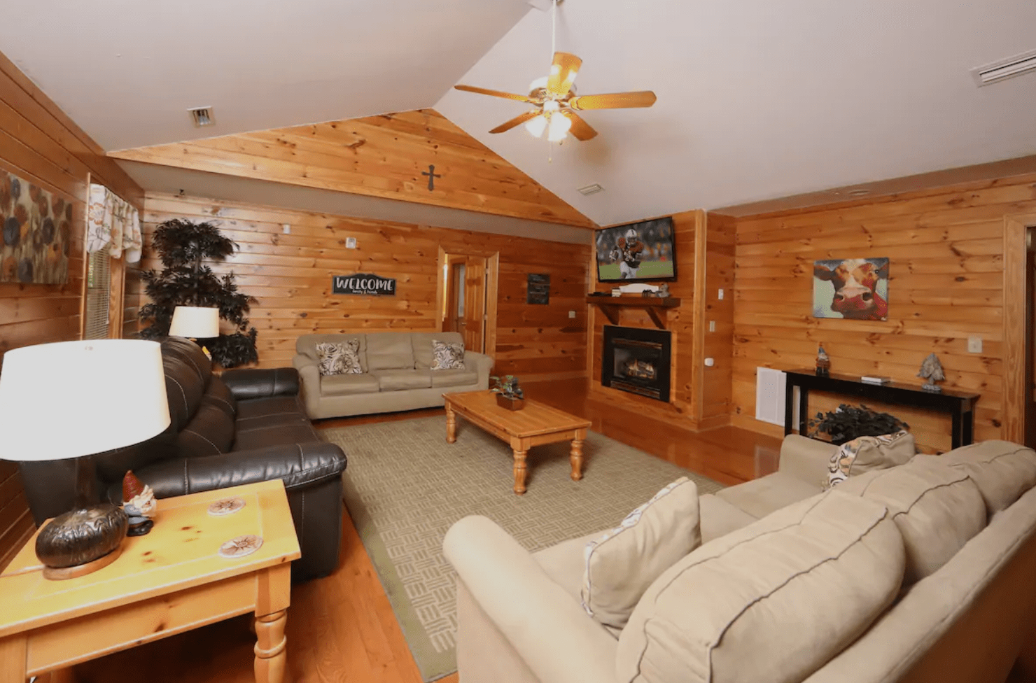 Sofas face a TV above a fireplace in a wooden living room. "WELCOME" sign on wall, ceiling fan overhead, decorative elements present.