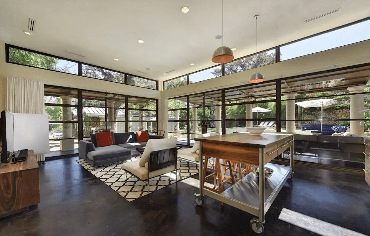 Seating arrangement with chairs and sofas faces outside large windows. Indoor island table on wheels under pendant lights. Lush greenery visible through glass walls.