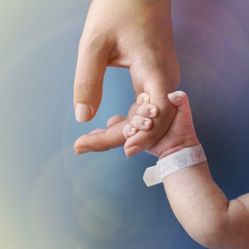 Baby hand grasps adult finger gently, against soft blue background. A hospital bracelet with text wraps the baby's wrist.