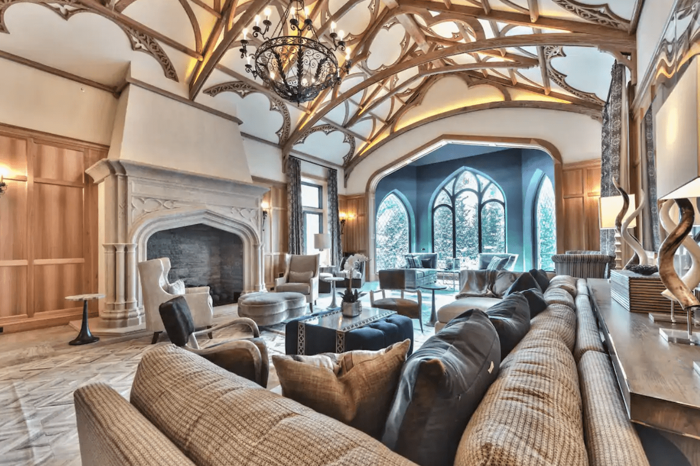 Elegant living room features ornate ceiling beams, a large stone fireplace, and luxurious seating. Tall arched windows overlook a garden.