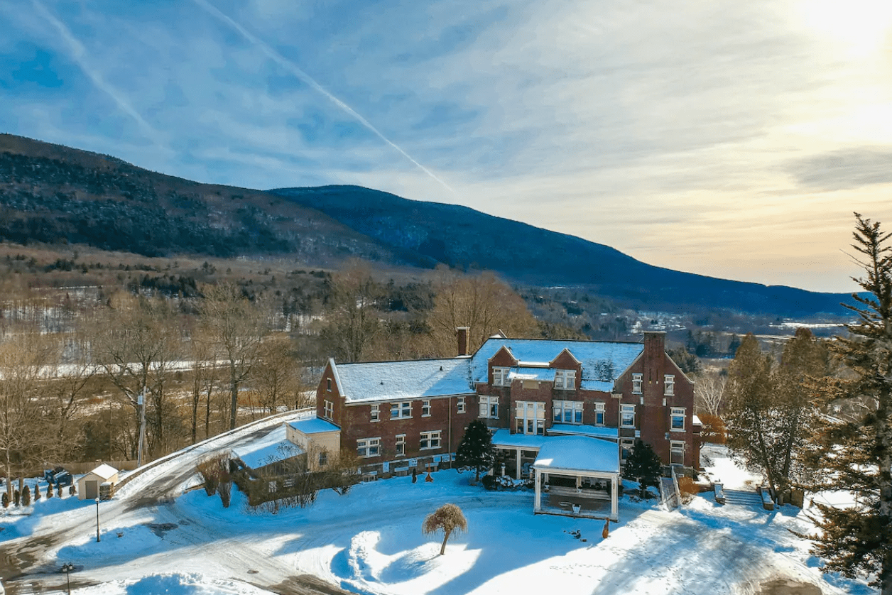 Red brick building covered in snow, set against a backdrop of rolling hills and a clear sky, surrounded by leafless winter trees.
