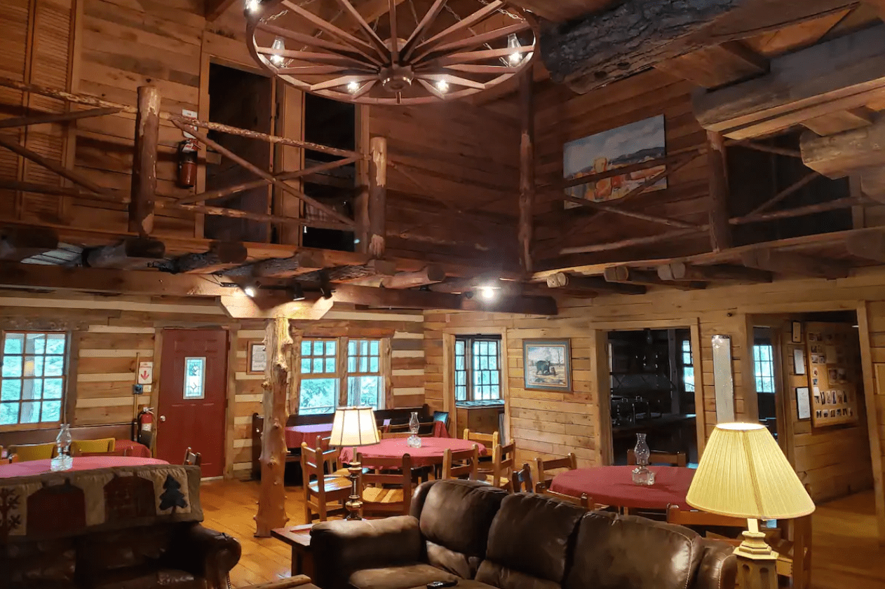 Rustic wooden interior features tables with red cloths, cushioned chairs, wagon-wheel chandelier overhead; cozy lodge-like setting with framed pictures on walls.