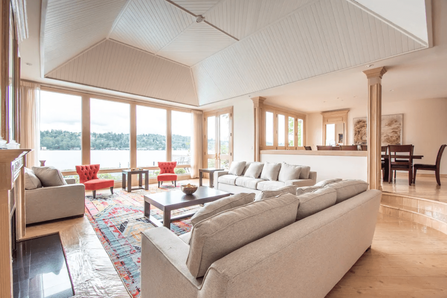 Beige sofas face windows, overlooking a lake. Red chairs accent the room. A patterned rug underlies coffee tables. Dining area visible in the background.