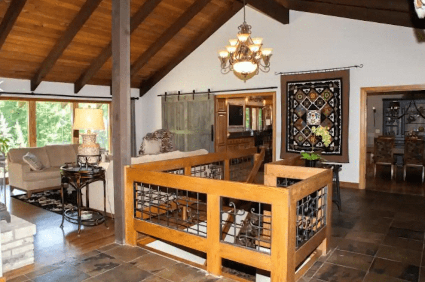 Wooden staircase railing overlooks living room, featuring a sofa and lamp, under vaulted ceiling with chandelier, surrounded by rustic decor and large windows.