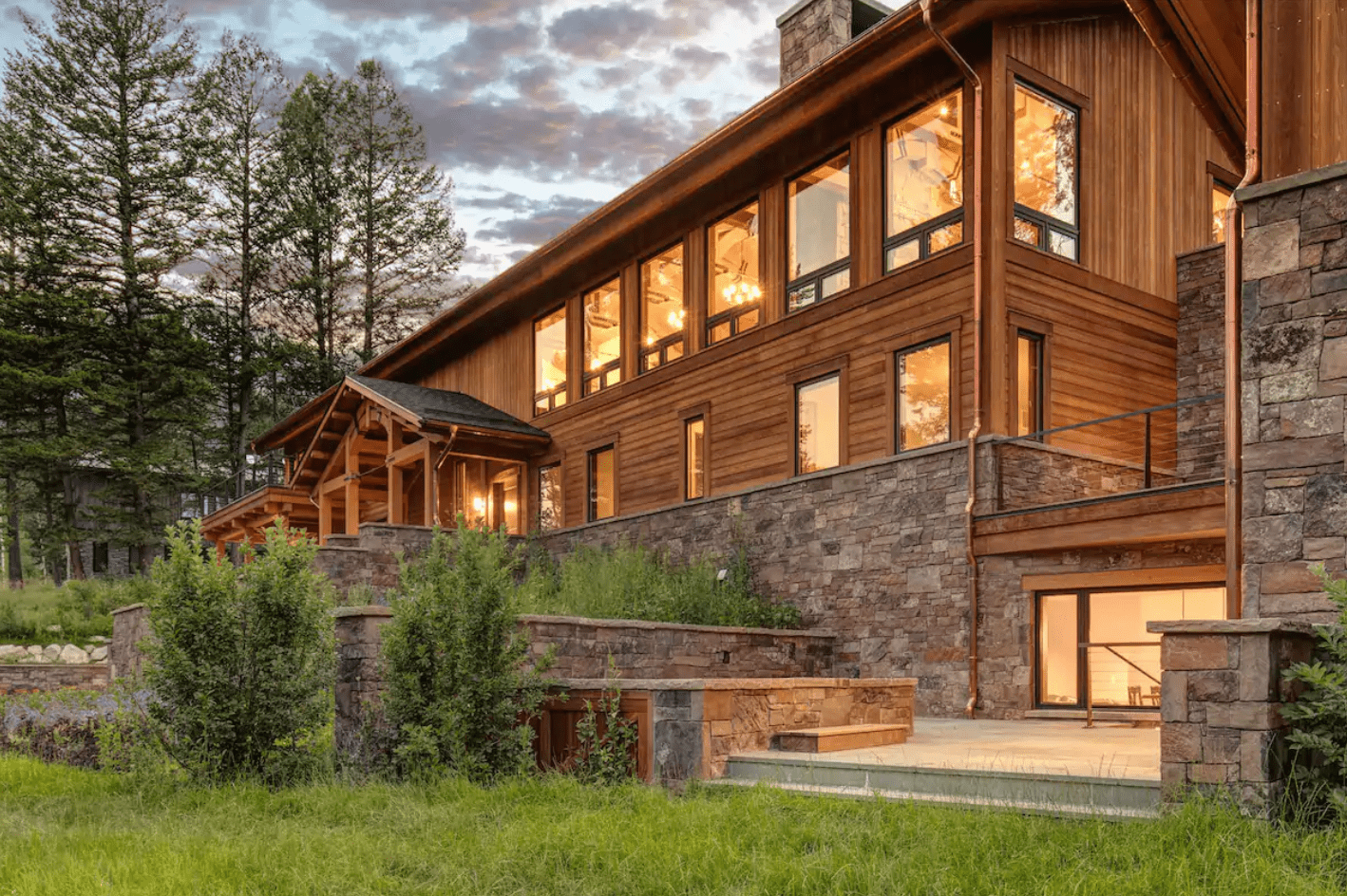 A large wooden house with large windows, surrounded by trees and grass, sits under a partly cloudy sky during sunset.