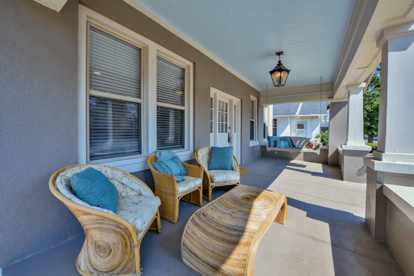 Rattan chairs with blue cushions rest on a covered porch, next to a swing and beneath a hanging light in a suburban setting.