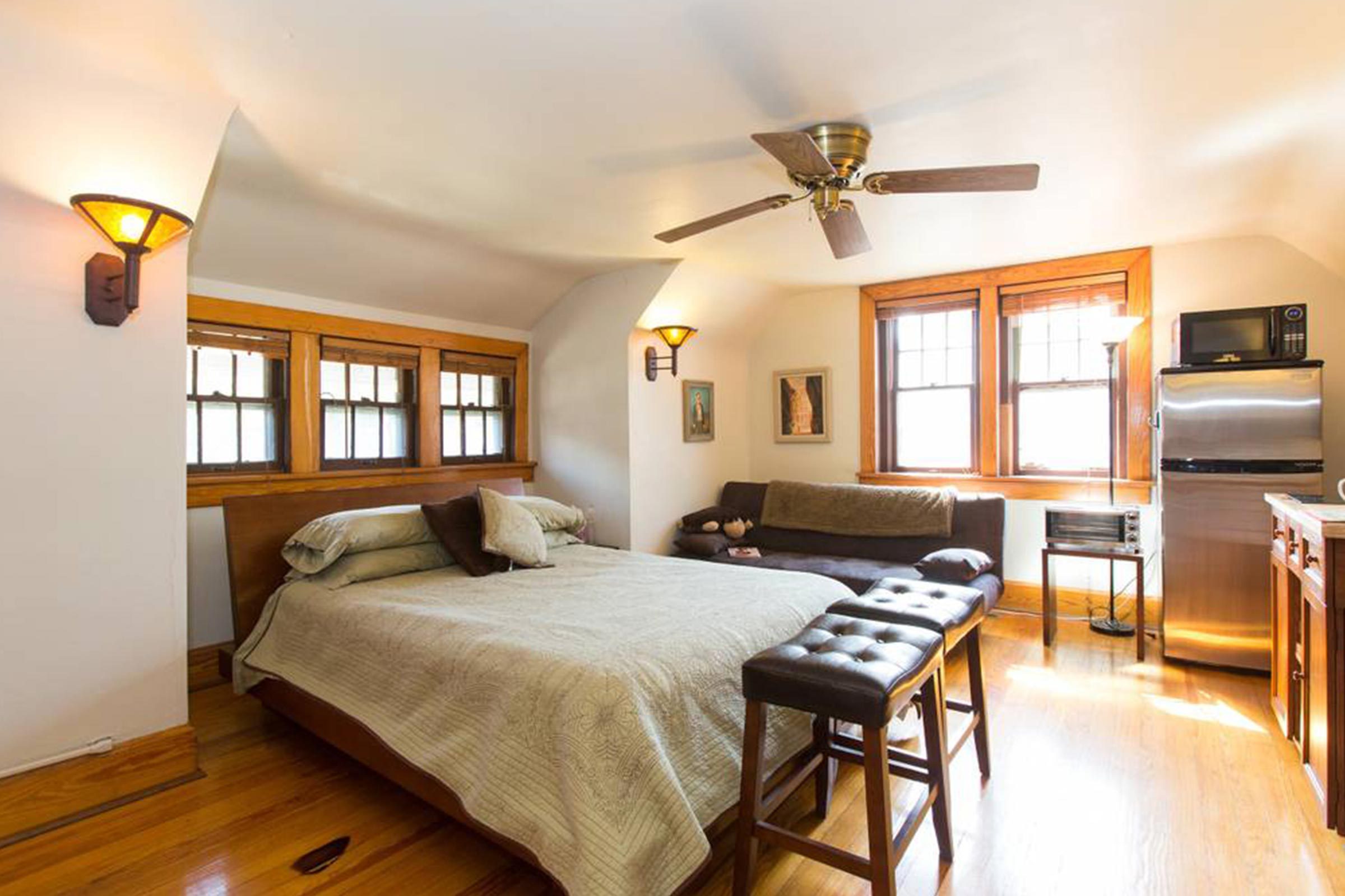 Bed sits under ceiling fan, surrounded by wooden furniture, modern appliances, and cozy decor in a sunlit room with multiple windows.