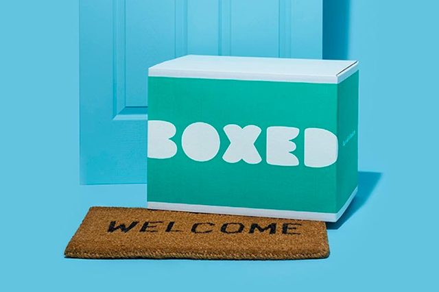 Box sits on doormat reading "WELCOME" in front of a blue door, against a blue wall background.