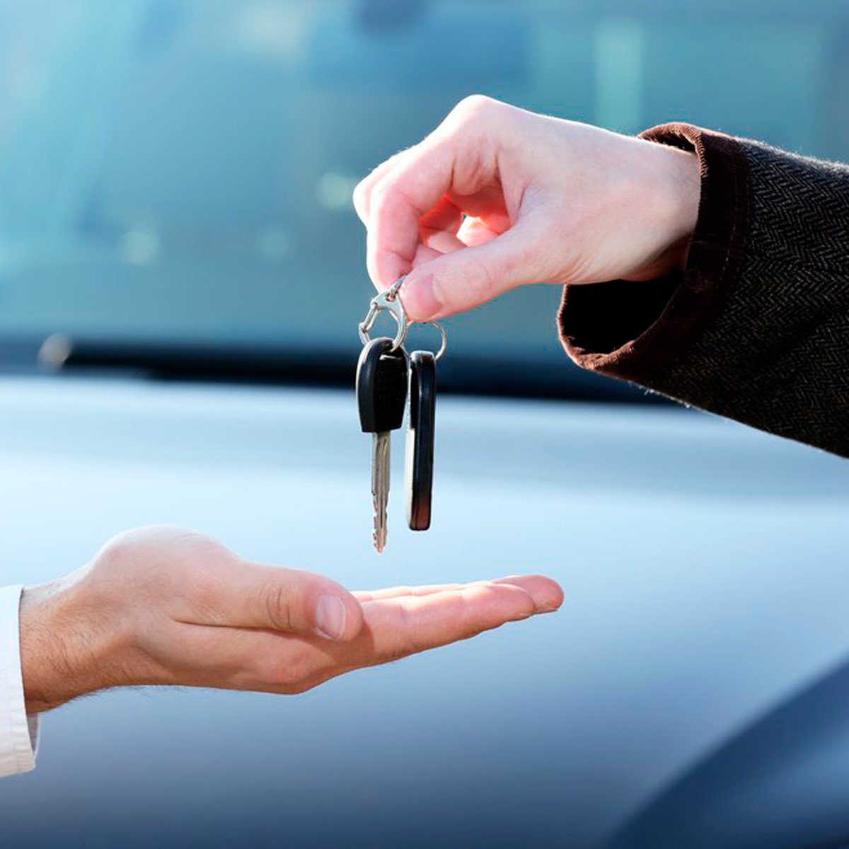 A hand holds car keys, passing them to an open palm, with a blurred vehicle in the background.