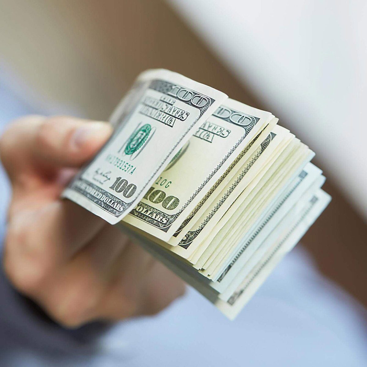 A hand holds a thick stack of hundred-dollar bills indoors, slightly blurred background.