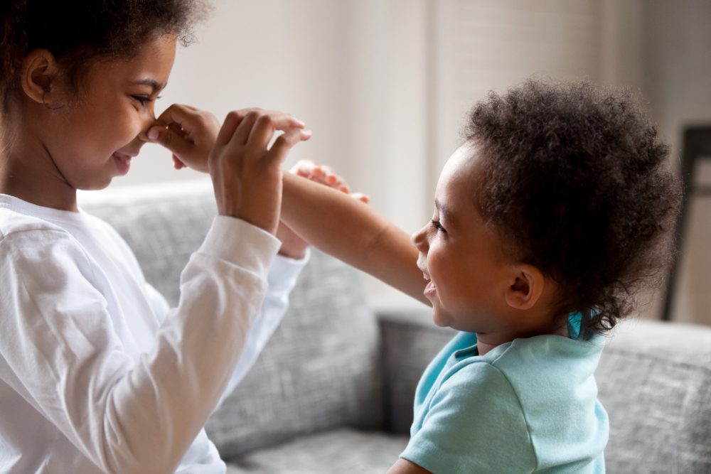 Close up Funny black children sitting on couch, little toddler adorable brother grabbed touch nose his elder small preschool lovely sister, two kids having fun together playing at home feeling happy