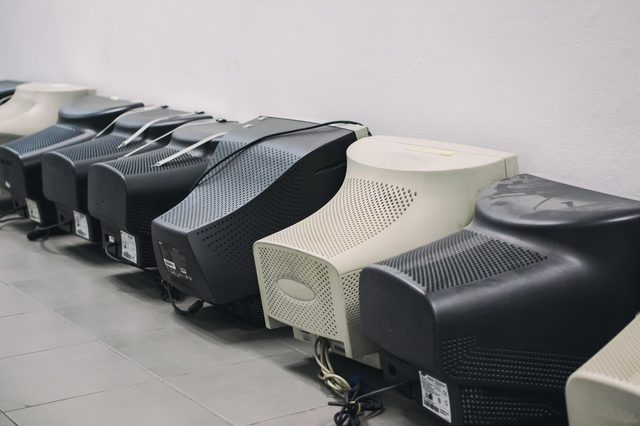 Obsolete CRT monitors sit in a row on tiled floor near a plain white wall, suggesting storage or disposal.