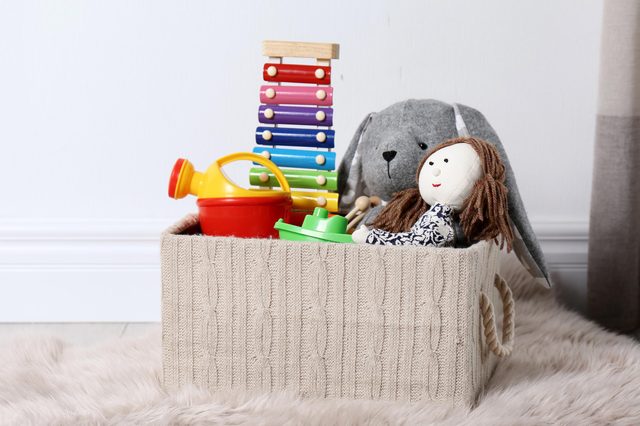 A knitted basket holds toys, including a xylophone, watering can, doll, and stuffed animal, on a fluffy rug beside a white wall.