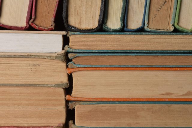 Books stacked horizontally, showing worn and colorful spines; packed tightly on a shelf in an indoor setting.