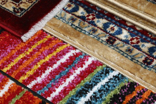 Colorful rugs display intricate patterns in bright stripes and traditional designs, overlapping in a vibrant, textured arrangement on the floor.