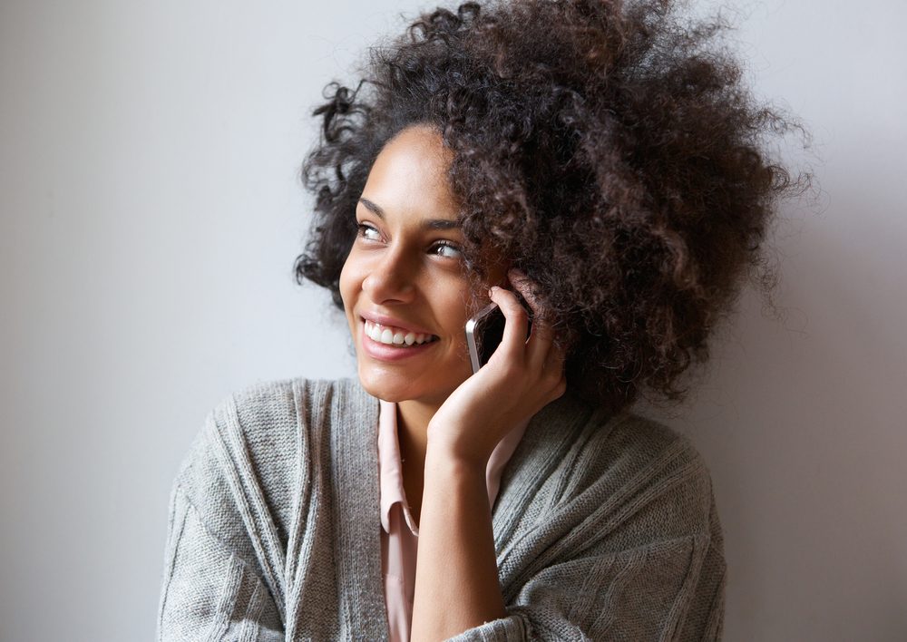 A person holds a phone to their ear, smiling, wearing a cozy sweater in a softly lit room.