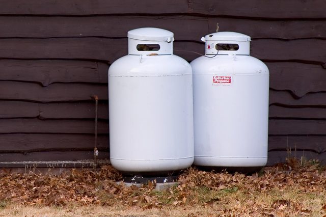 Two white propane tanks stand outdoors against a wooden wall, surrounded by fallen leaves and grass. One tank reads "Suburban Propane."