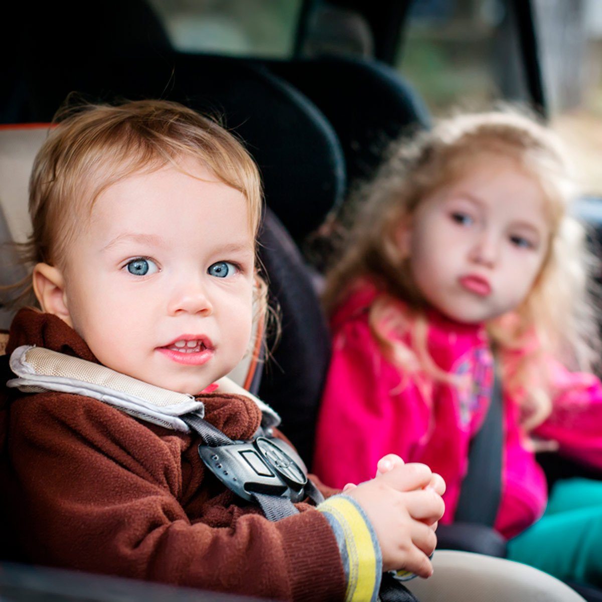 Two young children sit in car seats, looking forward; the background is the interior of a vehicle.