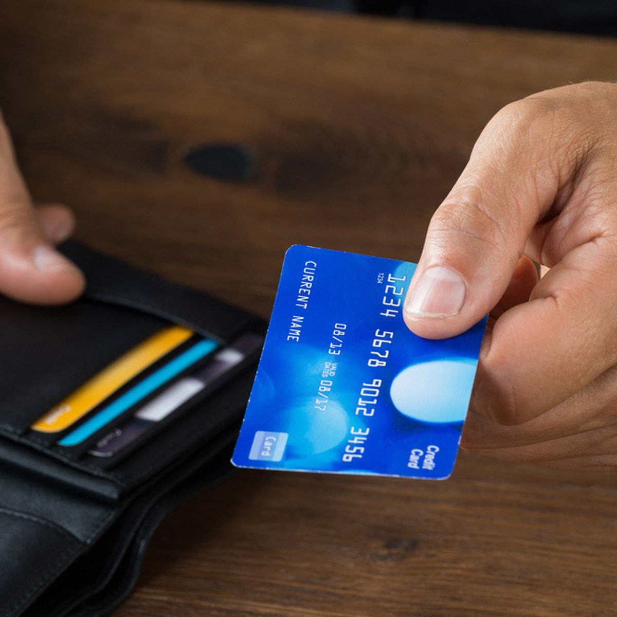 A hand holds a blue credit card near a black wallet on a wooden surface.
