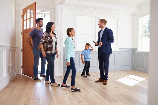 Adults and children enter an empty, sunlit room, meeting a man holding a tablet.