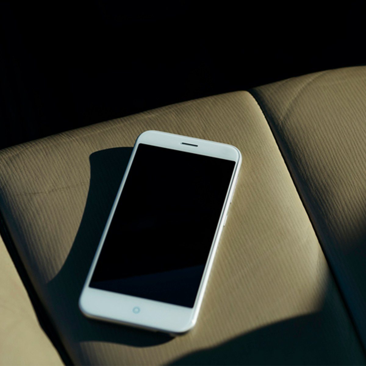 A smartphone rests motionless on beige car seat, partially bathed in sunlight.