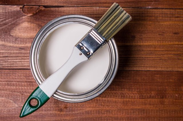 A paintbrush rests on an open can of white paint on a wooden surface.