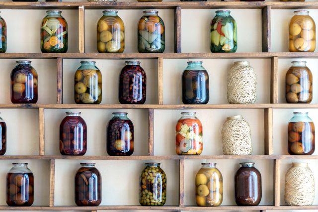 Jars filled with various pickled vegetables and fruits are displayed in a wooden shelf, creating a colorful and orderly arrangement against a plain wall.