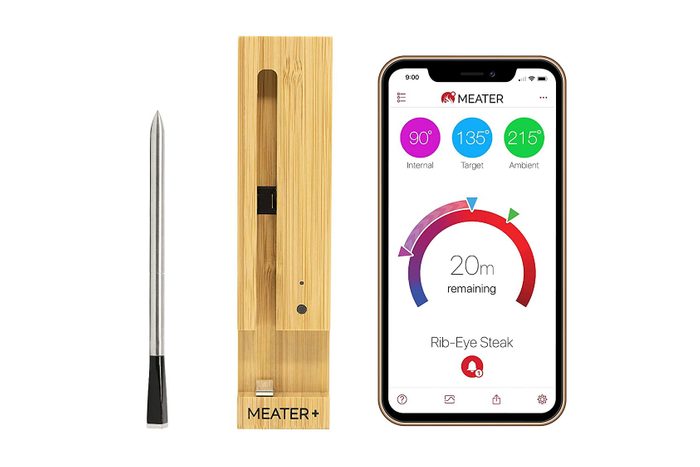 Meat thermometer displays temperature data on a smartphone app, with a wooden stand nearby. Text shows internal, target, and ambient temperatures for rib-eye steak.