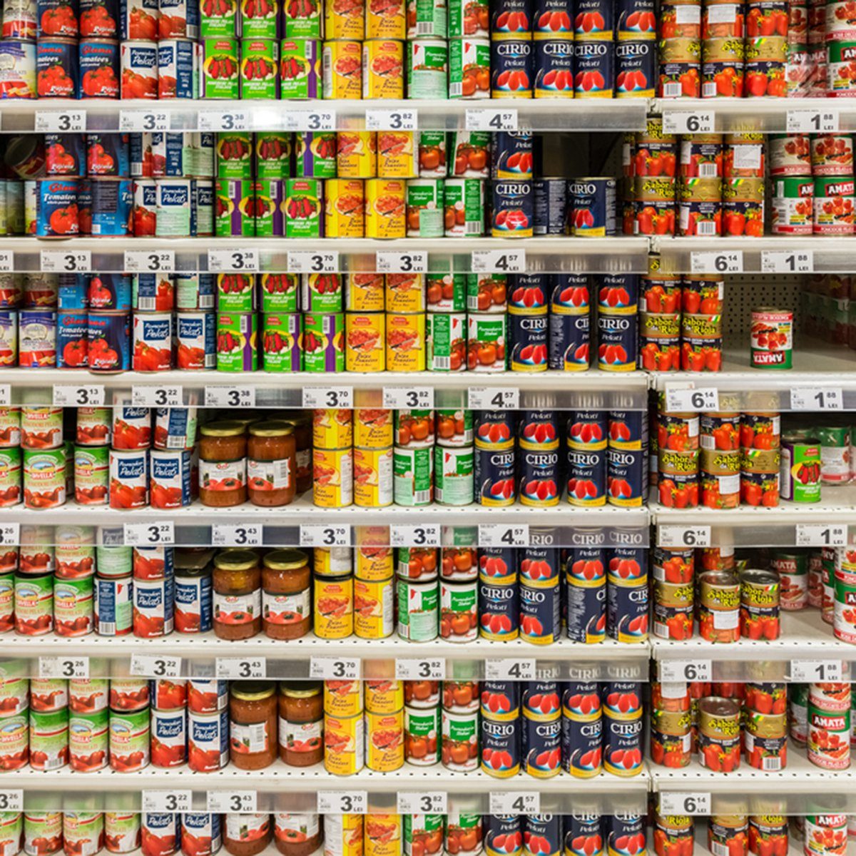 Cans of tomatoes stacked on supermarket shelves, sorted by brand and price, with labels displaying different product names and prices prominently visible.