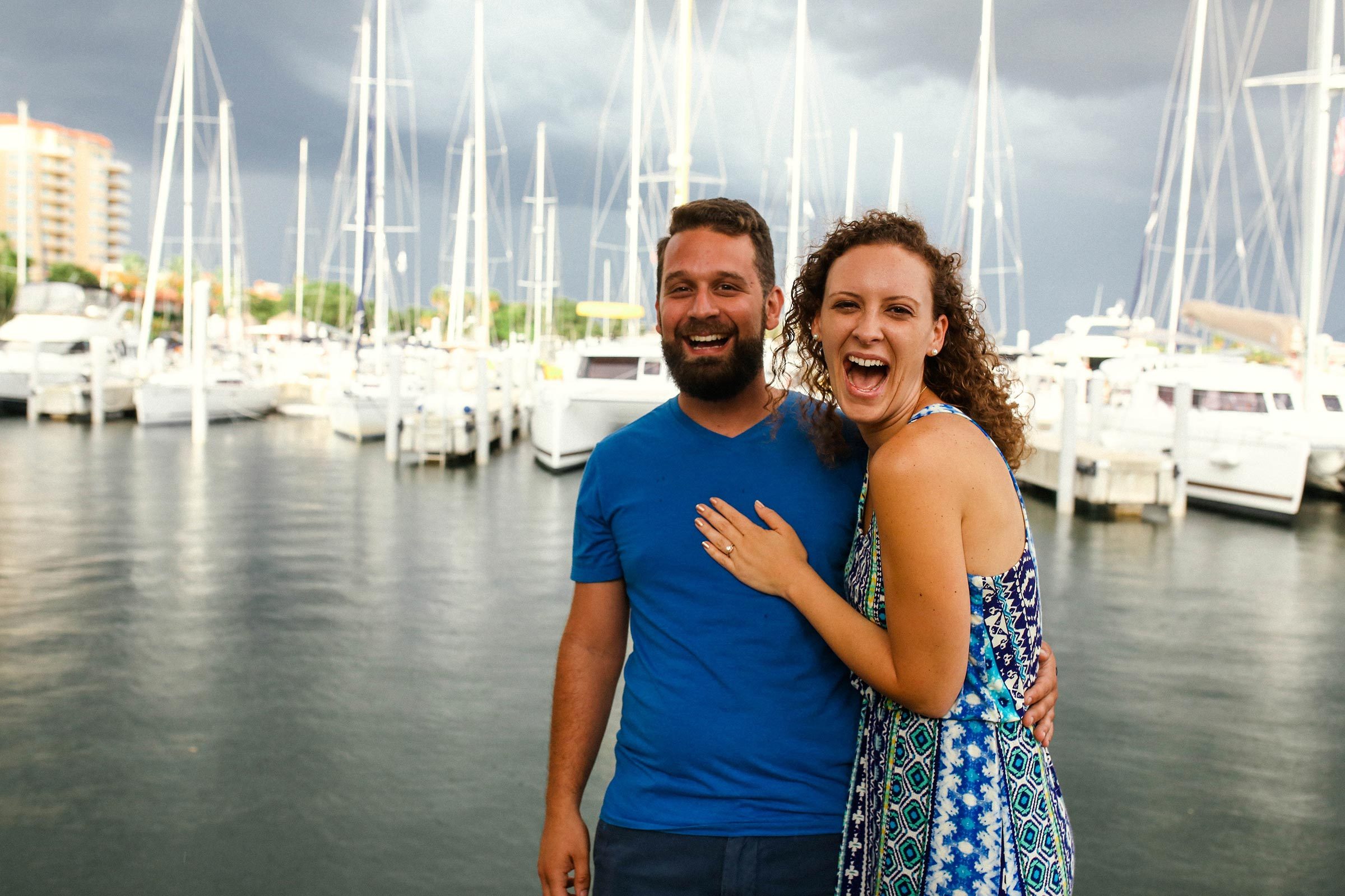 Two people smile joyfully, standing embraced. They're by a marina with numerous sailboats, under a cloudy sky.