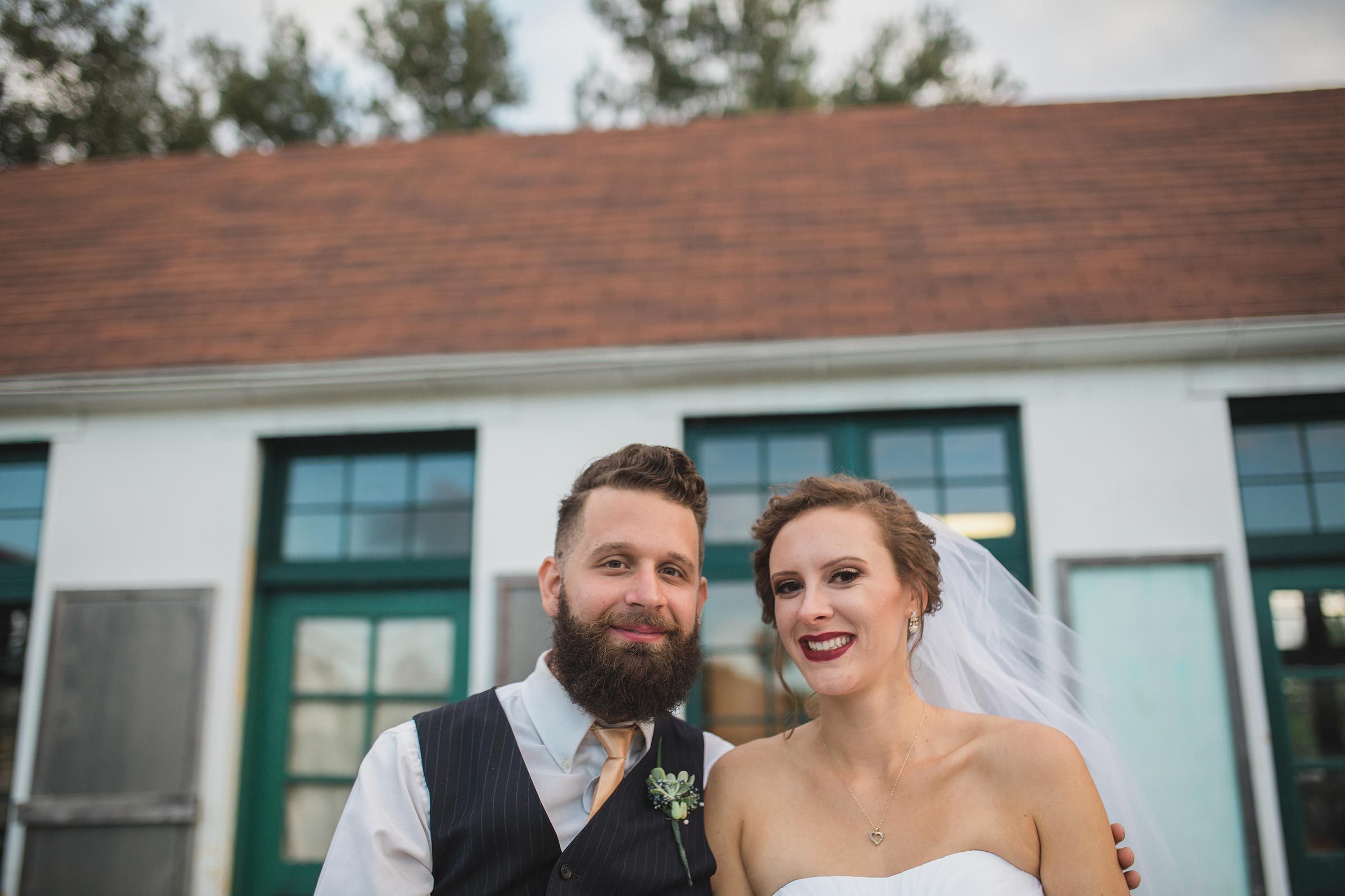 A smiling couple stands together outside, dressed in wedding attire, in front of a building with green-framed windows and a sloped roof.