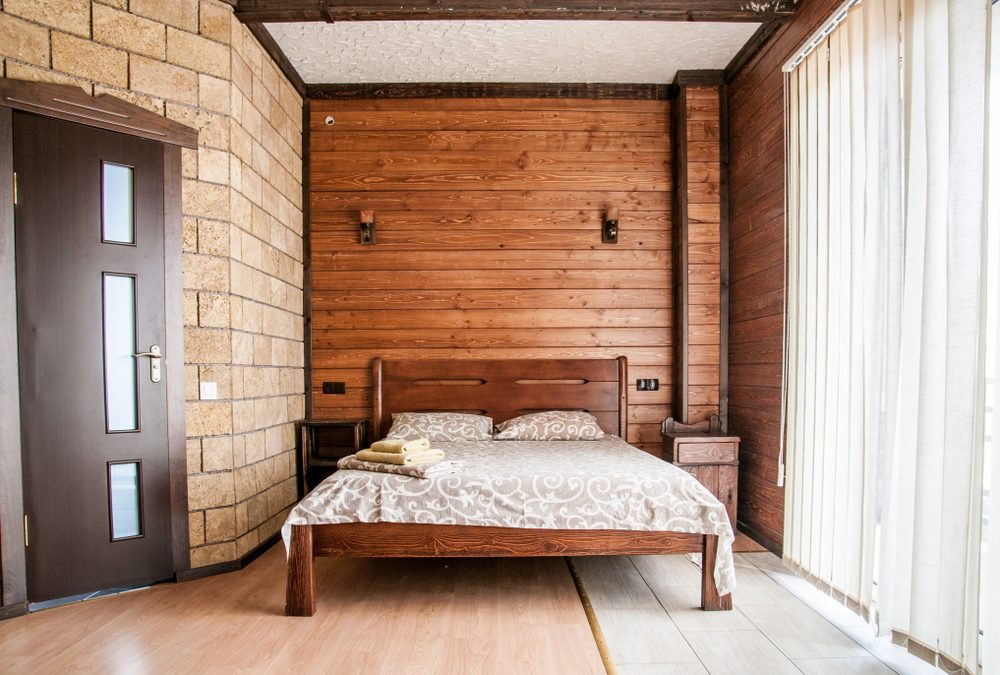 Bed with patterned comforter and folded towels; set in a wooden-walled room with large windows and vertical blinds.