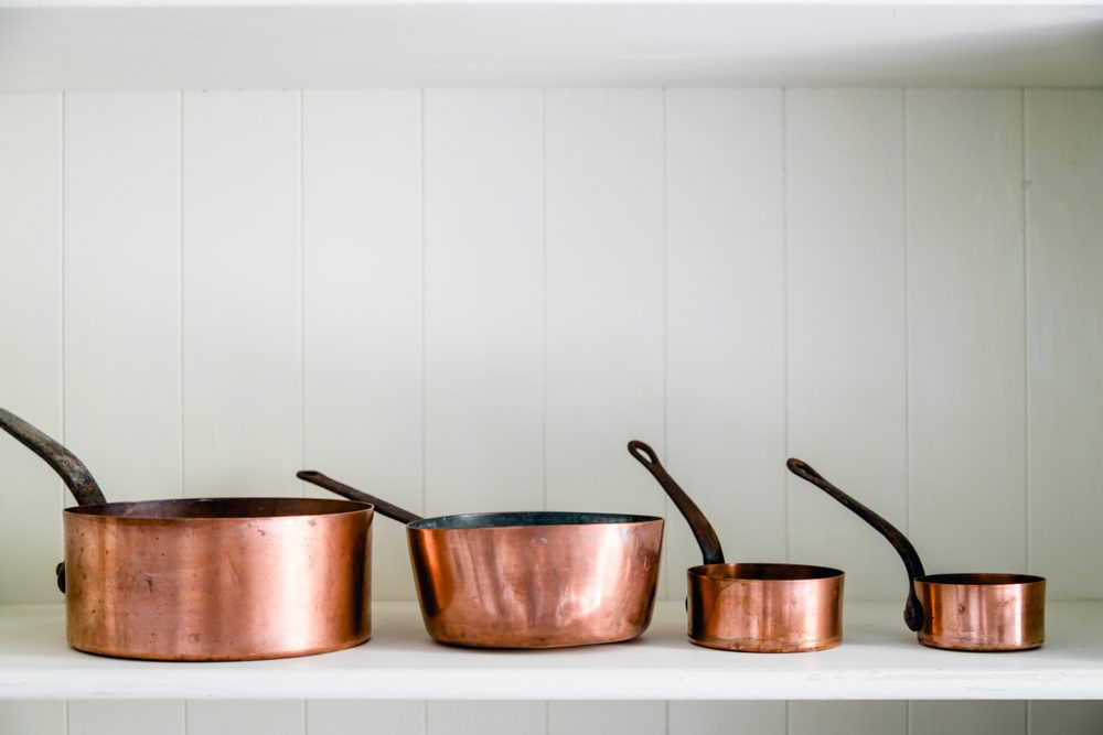 Copper pots are lined up on a white wooden shelf, each decreasing in size left to right, against vertical paneling.