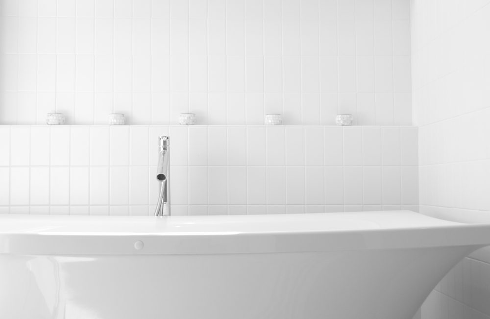 Bathtub sits beneath a shiny chrome faucet, surrounded by white tiled walls with small decorative jars lined up.