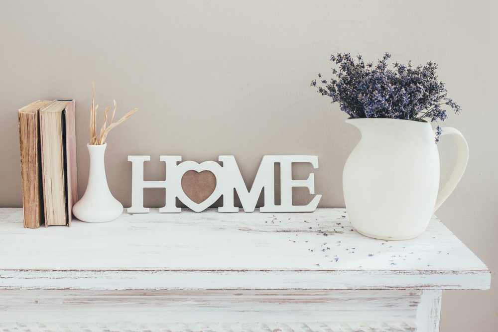 Books stand upright, with dried flowers and "HOME" sign in white on shelf; jug holds purple flowers on the right.