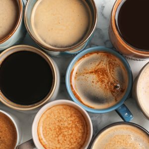 Various mugs contain diverse coffee types, arranged closely on a marble surface, showcasing different shades and textures of coffee and foam.