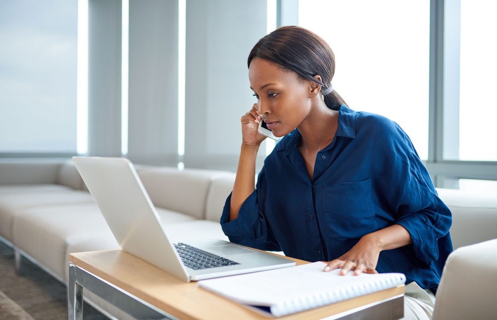 Person working intently on a laptop in a modern office, with a notebook on a sleek table and large windows in the background.