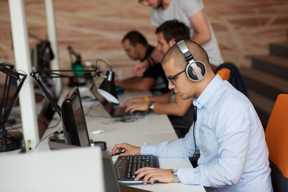 Individuals wearing headphones work on laptops in a modern office setting, focusing intently on screens at desks with lamps.