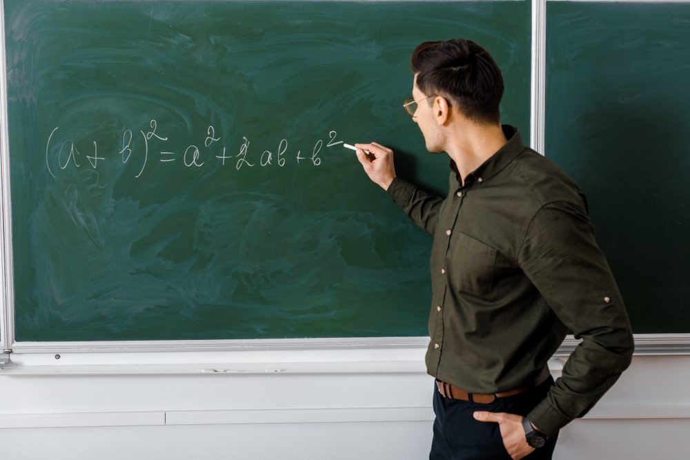 Man writing math equation on a green chalkboard in a classroom. Equation: (a + b)Â² = aÂ² + 2ab + bÂ².