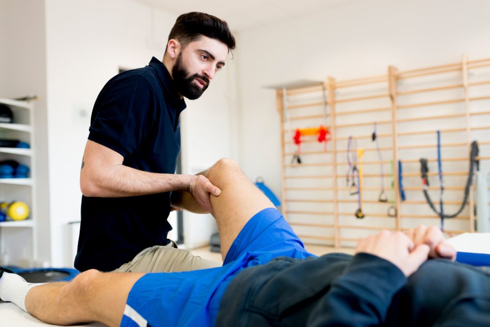 Therapist examines patient's knee, applying pressure, in a brightly lit physiotherapy room with exercise equipment on the wall.