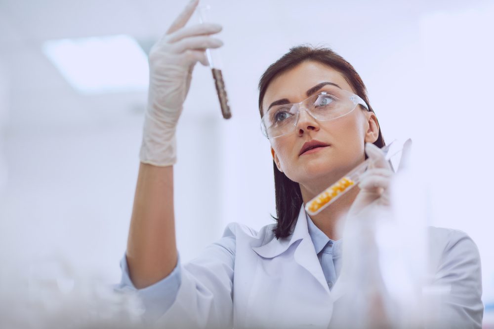 Scientist examines a test tube, wearing gloves and goggles, in a bright laboratory setting.