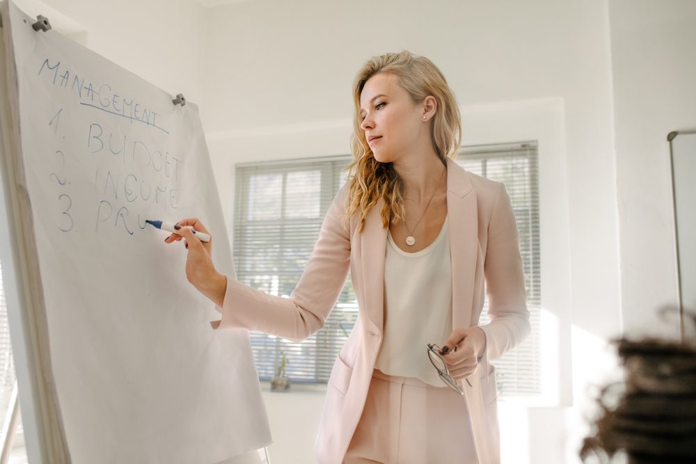A person writes on a flip chart listing "MANAGEMENT, BUDGET, INCOME, PRO" in a brightly lit office with large windows.