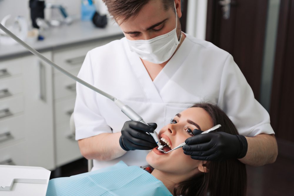 Dentist examines a patient's teeth, using dental tools, in a clinical setting with cabinets and equipment in the background.