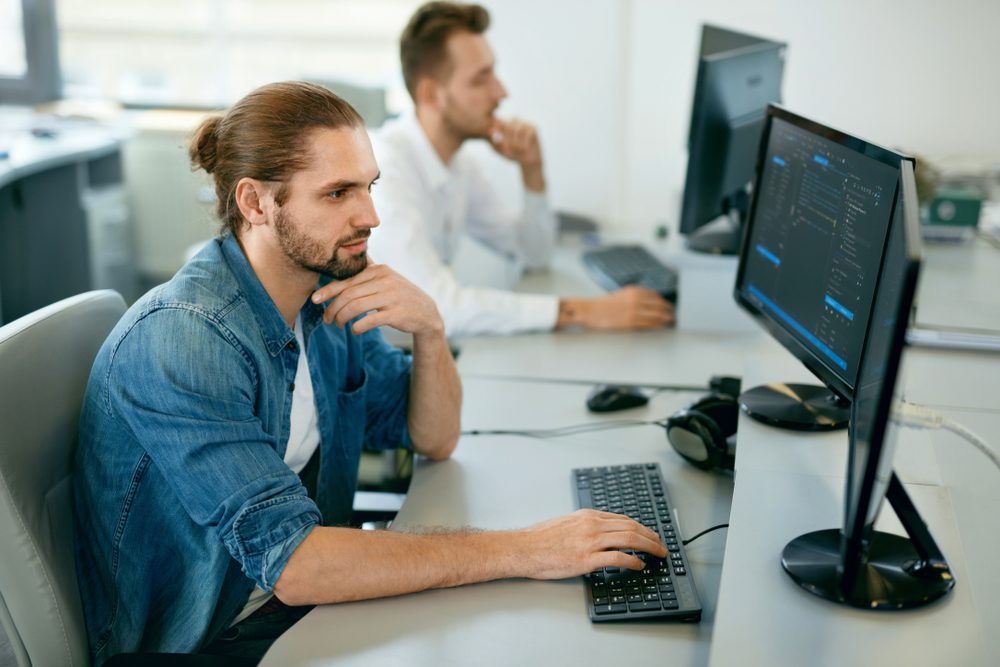 A man in a denim shirt types at a computer in an office, with another person working at a nearby desk.